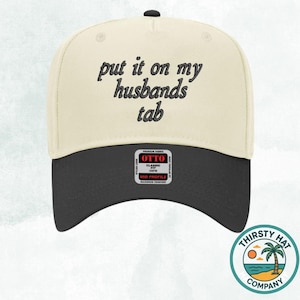 May include: A cream and black baseball cap with the text "put it on my husbands tab" in black script. The hat has a curved brim and a black button on top. A small tag reads "OTTO CLASSIC FIT". The Thirsty Hat Company logo is on the side.