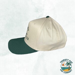 Puede incluir: Una gorra de b&eacute;isbol blanca y verde con el texto "The Country Club" bordado en la parte delantera. La gorra tiene una visera verde y una corona blanca.