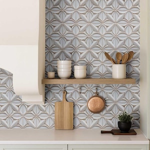 May include: A kitchen scene featuring a backsplash of patterned, light blue and white tiles. A wooden shelf holds white bowls and a ceramic utensil holder with wooden spoons. A wooden cutting board and copper pan hang below.