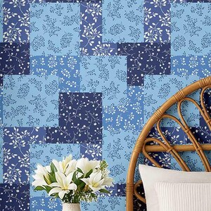 以下が含まれることがあります： 青色のパッチワーク柄の壁紙。白いアクセントのフローラルデザインが特徴です。白いユリの花束が花瓶に入って壁紙の前に置かれています。籐のヘッドボードと白い枕が背景にあります。