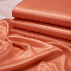 May include: Close-up of folded peach-coloured satin fabric. The fabric has a smooth, lustrous surface with visible folds and creases. The top layer is neatly folded, while the bottom layer is draped and flowing.