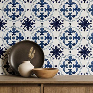 May include: A still life arrangement featuring a dark brown plate, a small white vase with dried grass, and a wooden bowl. The background showcases a repeating blue and white floral tile pattern, creating a decorative and artistic composition.