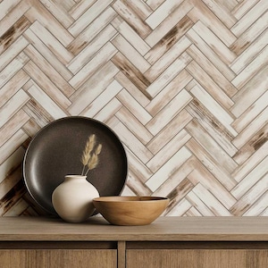 Puede incluir: Una composición decorativa con un plato marrón oscuro, un pequeño jarrón blanco con tallos secos y un cuenco de madera. El fondo es un patrón de espiga de tablones de madera claros y oscuros, creando una estética rústica. Los objetos se exhiben sobre una superficie de madera.