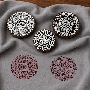 Puede incluir: Tres bloques de impresión circulares de madera marrón oscuro con intrincados patrones de mandala tallados en blanco, junto a dos impresiones circulares de color granate sobre una tela gris claro suave.