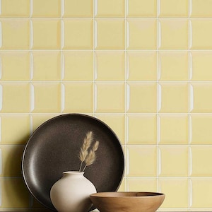 Peut inclure: Nature morte avec une assiette marron foncé, un petit vase blanc contenant de l'herbe séchée et un bol en bois. En arrière-plan, un mur de carreaux carrés jaune pâle avec des joints blancs, créant un motif géométrique texturé.