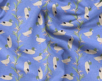 Puede incluir: Tela azul con un patrón repetido de patos blancos con detalles negros y cabezas verdes. Tallos florales verdes y beige se intercalan con los patos. La tela tiene una textura ligeramente arrugada.