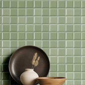 Pode incluir: Uma composição de natureza morta com um fundo de azulejos quadrados verde sálvia. Um prato castanho escuro, um pequeno vaso branco com espigas de trigo secas e uma tigela de madeira estão em primeiro plano. A cena evoca uma sensação de elegância rústica.