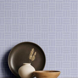 May include: A still life arrangement featuring a dark brown plate, a small white vase with dried wheat, and a wooden bowl. The background is a light purple tile pattern with white lines.
