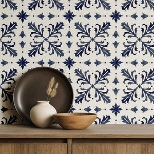 May include: A still-life arrangement featuring a dark brown plate, a small white vase with dried grass, and a wooden bowl on a wooden surface. The background showcases a white tile pattern with blue floral designs, creating a harmonious blend of textures and colors.