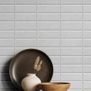 May include: A still-life composition featuring a dark brown plate, a small beige vase with dried wheat, and a wooden bowl. The background is a wall of white rectangular tiles, creating a minimalist aesthetic. The objects are arranged to create a sense of calm and simplicity.