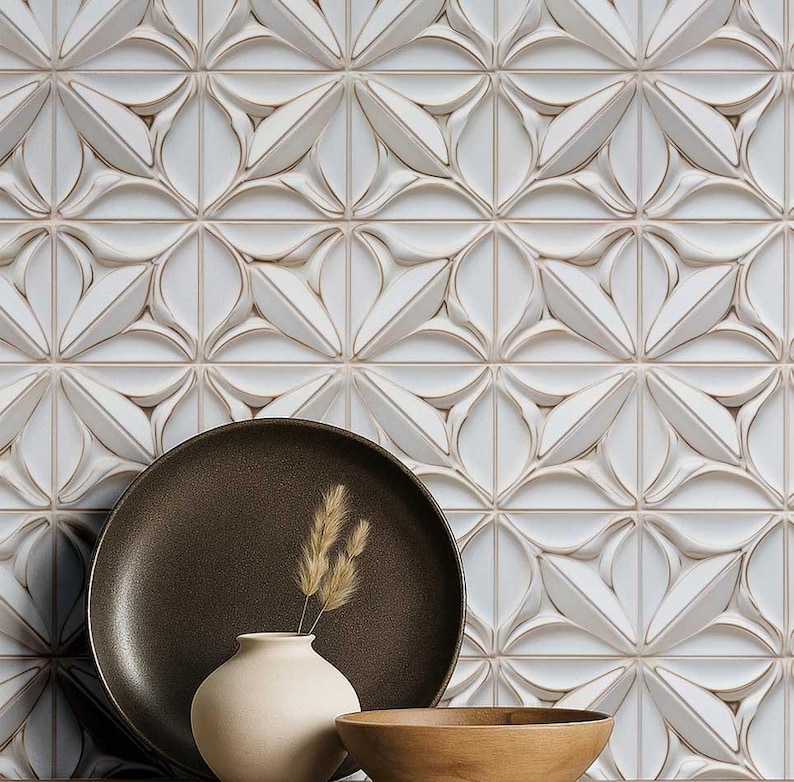 May include: A decorative arrangement featuring a white, textured tile backdrop with a repeating floral pattern. A dark brown plate, a small beige vase with dried wheat, and a wooden bowl are in the foreground. The scene evokes a sense of modern elegance.