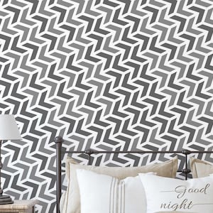 May include: A geometric pattern wallpaper in shades of gray and white, featuring a repeating chevron design. The wallpaper covers the wall behind a bed with a metal frame, pillows, and a nightstand with a lamp.