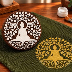 May include: A circular dark brown wooden stamp featuring a carved white silhouette of a Buddha figure sitting in a lotus position under a Bodhi tree with roots, placed next to a green fabric cloth with a gold stamped print of the same design.