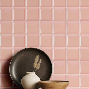 May include: A display of pink square tiles with a glossy finish. A dark brown plate, a white vase with dried grass, and a wooden bowl are in front of the tiles.