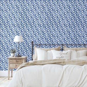 May include: A bedroom scene featuring a bed with white bedding and pillows. The wall is covered in a blue and white chevron pattern wallpaper. A nightstand with a lamp and a small plant is next to the bed.