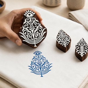 May include: Three dark brown wooden printing blocks with intricate floral carvings. One large block is held, while two smaller blocks sit on a white fabric surface. A blue ink floral pattern is stamped onto the fabric, matching the carved designs.