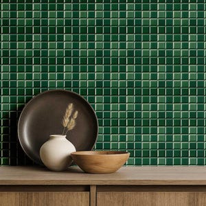 Puede incluir: Un bodegón con un plato marrón oscuro, un pequeño jarrón blanco con plantas secas y un cuenco de madera sobre una superficie de madera. El fondo es una pared de baldosas verdes esmeralda cuadradas y brillantes.