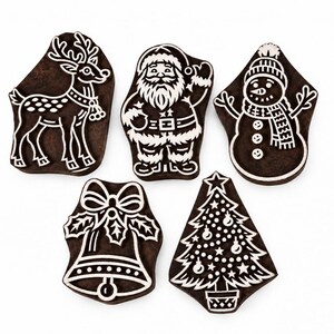 May include: A set of five dark brown wooden printing blocks featuring carved holiday designs including a reindeer, Santa Claus, a snowman, a bell with holly, and a decorated Christmas tree with a star on top, all with white outlines.