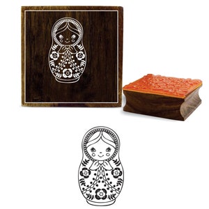 May include: Wooden stamp with a black and white illustration of a traditional Russian nesting doll, or matryoshka doll, with a floral pattern. The stamp is mounted on a wooden block.