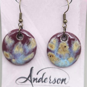 May include: A pair of round ceramic earrings with a burgundy base color and blue and gold accents. The earrings have bronze-colored hooks. The Anderson Stoneware logo is visible on the backing card.