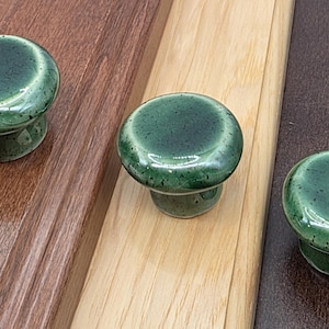 May include: Three round ceramic cabinet knobs with a glossy, speckled forest green glaze. Each knob features a smooth, domed top and a tapered base, showcasing a reflective finish with subtle dark green and black flecks throughout the surface.