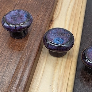May include: Three round cabinet knobs with a glossy, speckled finish in shades of deep purple, blue, and black. Each knob features a smooth, domed surface and is mounted onto a wooden panel, showcasing a distinct color gradient and reflective glaze.