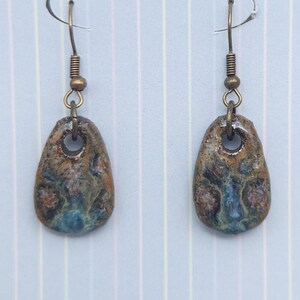 May include: A pair of handcrafted earrings with a unique, organic design. Each earring features a teardrop-shaped, textured stone with a blend of brown, blue, and tan hues. The earrings are suspended from bronze-colored ear wires.