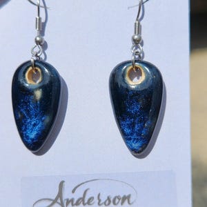 May include: A pair of handcrafted, teardrop-shaped earrings. The earrings feature a dark blue, glossy finish with a unique, textured blue pattern. Each earring is suspended from a silver-colored hook. The brand name "Anderson Stoneware" is visible on the bottom.