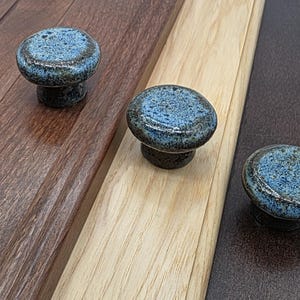 May include: Three round ceramic cabinet knobs with a speckled blue and dark brown glaze. Each knob features a smooth, rounded top surface and a tapered base, mounted individually on three distinct wooden panels of varying wood tones.