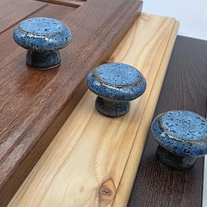 May include: Three blue ceramic cabinet knobs with a speckled glaze and a dark rim. The knobs are mushroom-shaped and sit on wooden surfaces of varying colors, including dark brown, light brown, and medium brown.
