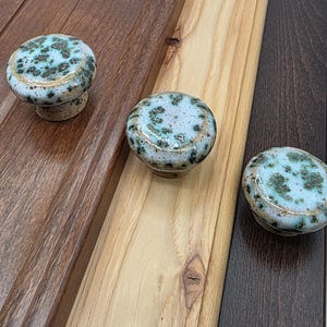 May include: Three ceramic cabinet knobs with a speckled blue and green design. The knobs have a light beige base and a gold-colored rim. The knobs are displayed on three different wood surfaces.