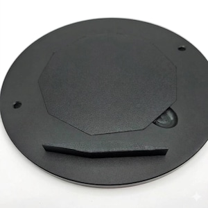 May include: A black, circular product base with a textured surface. The base features an octagonal inset and a raised rectangular section. Two small holes are visible on the edge. The base is designed for supporting another object.