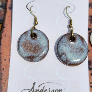 May include: A pair of handcrafted ceramic earrings. Each earring features a round, glazed pendant with a mottled blue and brown pattern. The earrings have brass-colored ear wires and a small hole at the top for attachment. The earrings are from Anderson Stoneware.