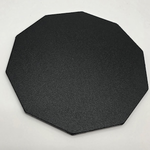 May include: A black, octagonal coaster with a textured surface. The coaster is made of a solid, dark material and is designed to protect surfaces from drink condensation and heat.
