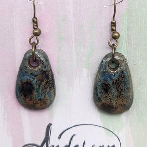 May include: A pair of handcrafted earrings with a unique, organic design. Each earring features a teardrop-shaped, glazed ceramic bead in shades of blue, brown, and black, suspended from antique brass ear wires. The earrings are from Anderson Stoneware.