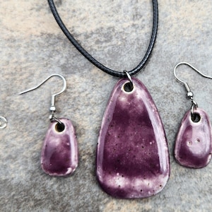 May include: A jewelry set featuring a pendant necklace and matching earrings. The pendant and earrings are a deep purple with a speckled texture and a glossy finish. The necklace has a black cord, and the earrings have silver-colored hooks.