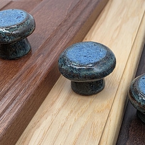 May include: Three ceramic cabinet knobs with a mushroom shape. The knobs have a blue and green speckled glaze and are mounted on wooden surfaces of varying colors: brown, light brown, and dark brown.