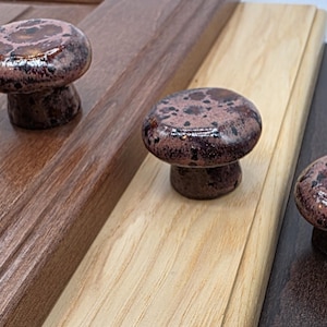 May include: Three mushroom-shaped cabinet knobs with a speckled, reddish-brown glaze. The knobs are displayed on wooden surfaces of varying colors: dark brown, light brown, and black. The knobs have a glossy finish.