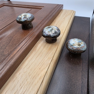 May include: Close-up of three ceramic cabinet knobs with a speckled, iridescent finish. The knobs are mounted on wooden panels in shades of brown and natural wood. The knobs have a mushroom shape.