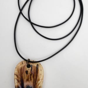 May include: A teardrop-shaped pendant necklace with a black cord. The pendant features a unique design with brown, beige, and blue hues. The necklace has a bronze-colored clasp.