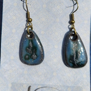May include: A pair of handcrafted earrings with a teardrop shape. The earrings feature a blue and green glazed surface with a speckled pattern. They have bronze-colored ear wires and a small loop at the top. The brand name "Anderson Stoneware" is visible.