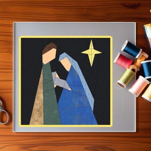 May include: A framed geometric art piece depicting a nativity scene with a yellow star on a black background. The artwork is surrounded by a yellow border and set on a wooden surface with scissors, spools of thread, and a small quilt.