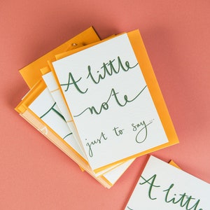 Peut inclure: Une pile de cartes blanches avec des enveloppes orange. Les cartes portent l'inscription en lettres vertes "A little note just to say..."