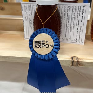 May include: A jar of honey with a white lid and a gold string, displayed on a wooden shelf. A blue ribbon with the text "BEE & EXPO" is in front of the jar. Judging criteria and comments are visible in the background.