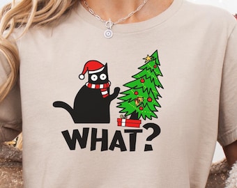 May include: Beige t-shirt with a graphic of a black cat wearing a Santa hat and scarf, next to a Christmas tree. The word "WHAT?" is printed below the cat. The person is wearing red and black plaid pajama pants.
