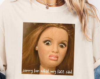 May include: Ivory-colored t-shirt featuring a square image of a doll's face with a surprised expression. The text "sorry for what my face said" is below the image. The shirt has rolled-up sleeves.