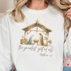 May include: White sweatshirt with a watercolour illustration of the Nativity scene. The image includes a stable, figures, and the text "the greatest gift of all" and "Matthew 1:2". The sweatshirt is worn by a person.