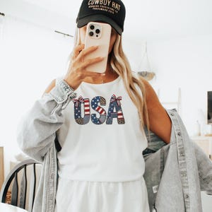 Peut inclure: Haut sans manches blanc avec le mot "USA" dans un design patriotique. Les lettres sont décorées de motifs du drapeau américain. Le modèle porte une casquette de baseball noire et un short blanc.
