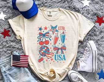 May include: Cream-colored t-shirt with a patriotic design featuring stars, bows, cherries, and the text "USA" and "1776". The shirt is paired with denim shorts, a blue and white trucker hat, and white sneakers.