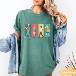 May include: Light green t-shirt featuring a graphic of four anime characters walking through colorful doors. The design includes characters in various outfits and poses. The shirt is a relaxed fit.